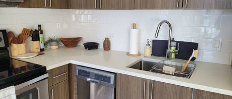Een moderne keuken met houten kasten, een roestvrijstalen vaatwasser, een spoelbak met afwassysteem en diverse keukenbenodigdheden en apparaten.