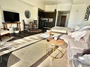 Een moderne woonkamer met een flatscreen televisie, een eethoek met een tafel en stoelen, een keuken op de achtergrond, en een slaapkamer met een bed en een slaapbank.