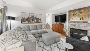 A modern living room with a grey sectional sofa, a grey ottoman, and a black armchair. There is a flat-screen TV mounted on the wall, a stone fireplace, and a framed sign above the fireplace. - Wi1073-2 (Keystone)