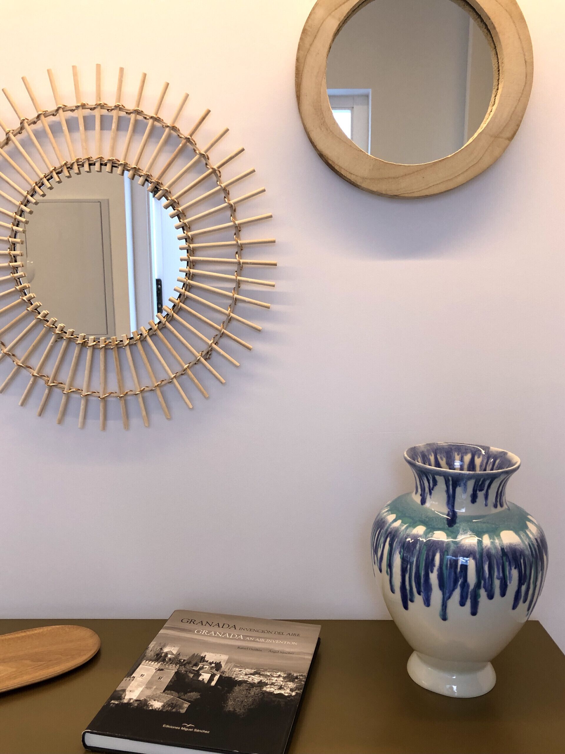 A room with a wooden wall hanging, a mirror, a vase, and a book on a table.