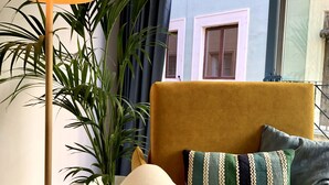 En hyggelig stue med en gul sofa, et hvidt pled og en pude med striber i grøn og hvid. Der er en høj gulvlampe med en beige skærm og en potteplante ved siden af.