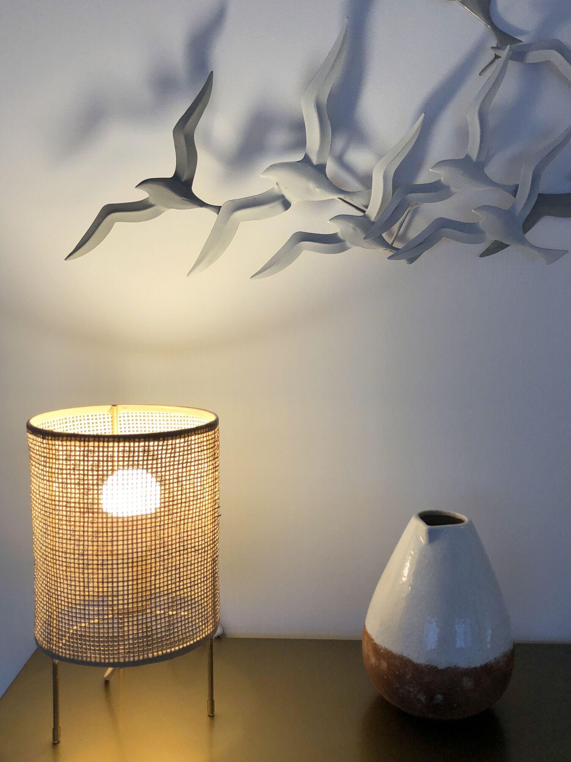Een lamp met een gevlochten kap en een vaas op een tafel, voor een muur met een vogelmuurdecoratie.