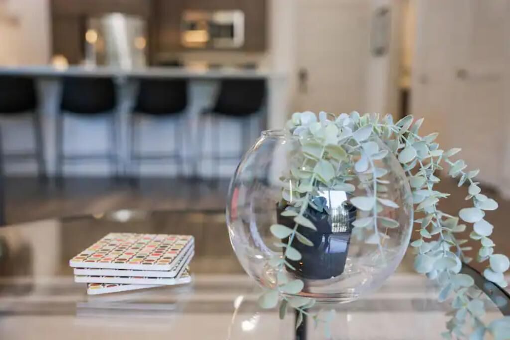 A clear glass vase with a plant on a reflective surface, a small stack of books, and a blurred background of a modern interior space.