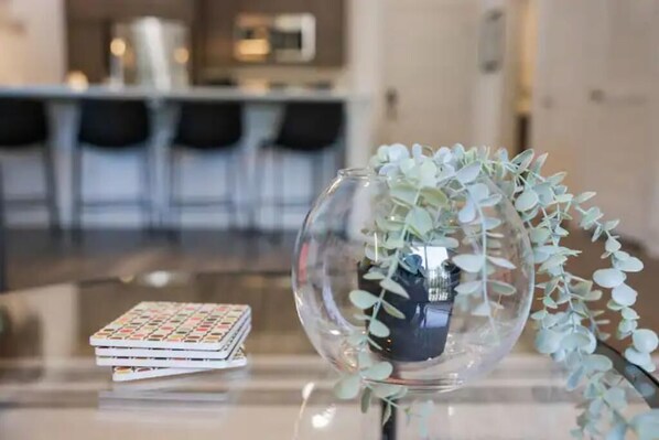 A clear glass vase with a plant on a reflective surface, a small stack of books, and a blurred background of a modern interior space.