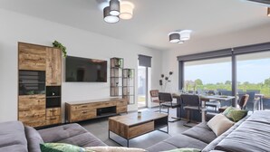Een moderne woonkamer met een grote flatscreen televisie, houten meubels, een eethoek met tafel en stoelen, en een balkon met uitzicht.