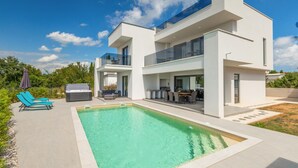 Een modern huis met twee verdiepingen, een zwembad, buitenzitplaatsen en een glazen hekwerk.