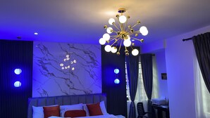 A modern hotel room with a marble wall, a large bed with red and white pillows, a unique chandelier, and dark curtains.