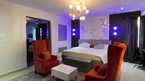 A modern hotel room with a large bed, two red armchairs, a white coffee table, and a unique chandelier.