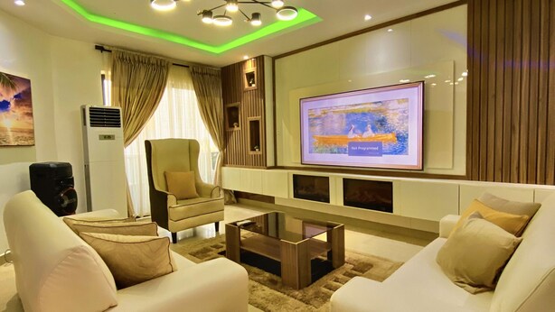 A modern living room with two white sofas, a coffee table, a flat-screen TV mounted on the wall, and a ceiling with green lighting accents.