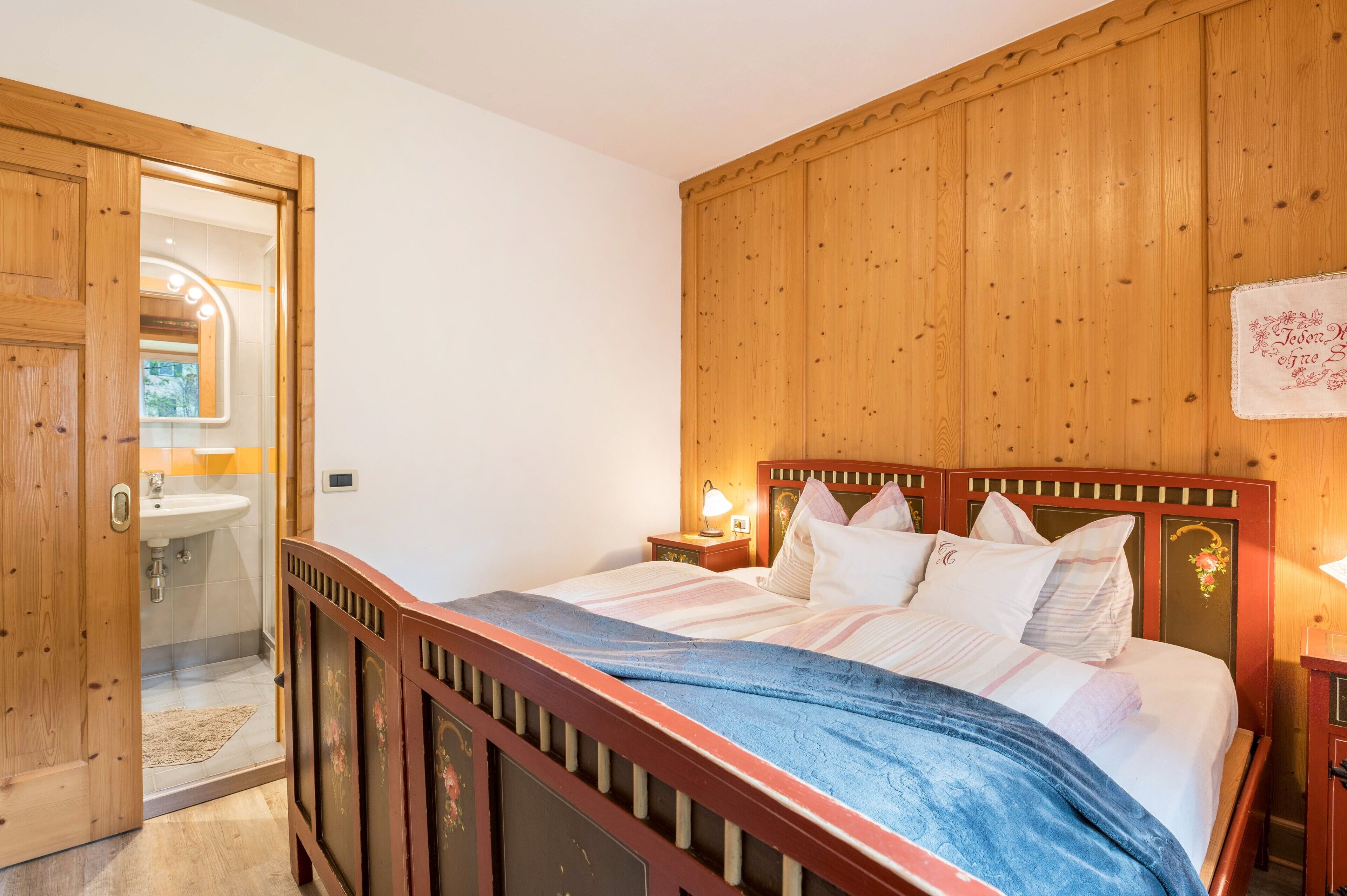 A bedroom with a wooden headboard, a bed with white bedding, a nightstand with a lamp, and a wooden door leading to a bathroom.