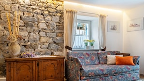 A room with a stone wall, a wooden dresser, a patterned sofa, and a window with curtains. - Apartment 'Ciasa Majonade Pozza Di Fassa Tipo1' with Mountain View, Private Terrace and Wi-Fi (Pozza di Fassa)