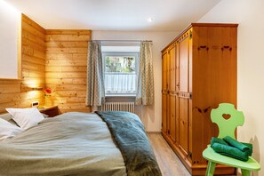 A bedroom with a wooden headboard, a bed with pillows, a nightstand with a lamp, a window with curtains, and a wooden wardrobe.