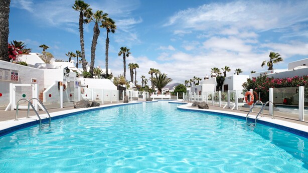 A swimming pool with clear blue water, surrounded by white buildings with palm trees and a clear sky. - Vacation Apartment 'Puerto Chico H' with Shared Pool, Private Terrace and Wi-Fi (Playa Blanca)