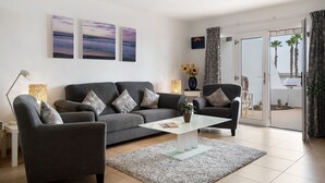 A modern living room with a grey sofa, a glass coffee table, and a view of palm trees through a sliding glass door. - Vacation Apartment 'Puerto Chico H' with Shared Pool, Private Terrace and Wi-Fi (Playa Blanca)