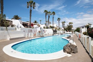 Une piscine bordée d’une terrasse blanche, des palmiers et des bâtiments blancs, le tout sous un ciel dégagé.