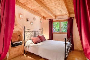 A bedroom with a wooden ceiling, a bed with a white bedspread, two pink pillows, a nightstand with a radio, and a window with red curtains.