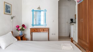 Ein Schlafzimmer mit einem Bett, einer Holzkommode, einem kleinen Tisch mit Blumen in einer Vase, einem spiegelnden Wandbild mit blauem Rahmen und einer weißen Tür, die zur Küche führt.