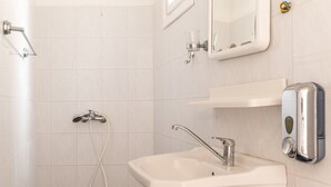 Un baño con un lavamanos blanco, un cabezal de ducha de mano, un espejo y un dispensador de jabón.