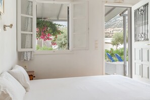 A bedroom with a bed, a window with curtains, and an open door leading to an outdoor area with a view of greenery and a mountain.