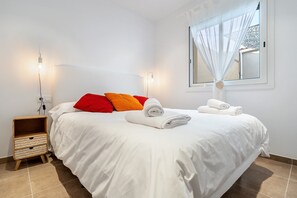 Ein ordentlich bezogenes Bett mit weißer Bettwäsche, roten und orangefarbenen Kissen und einem aufgerollten Handtuch an der Seite. Dazu ein kleiner Nachttisch mit einem gewebten Korb.