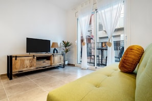Een woonkamer met een bank, een televisie op een houten kast, een plant en een balkon met een tafel en stoelen.