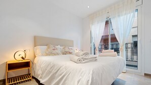 A neatly made bed with white linens, a bedside table with a lamp, and a view of a balcony through sheer curtains. - Apartment 'Mabicarent Playa 2' with Private Terrace, Wi-Fi and Air Conditioning (Calafell)