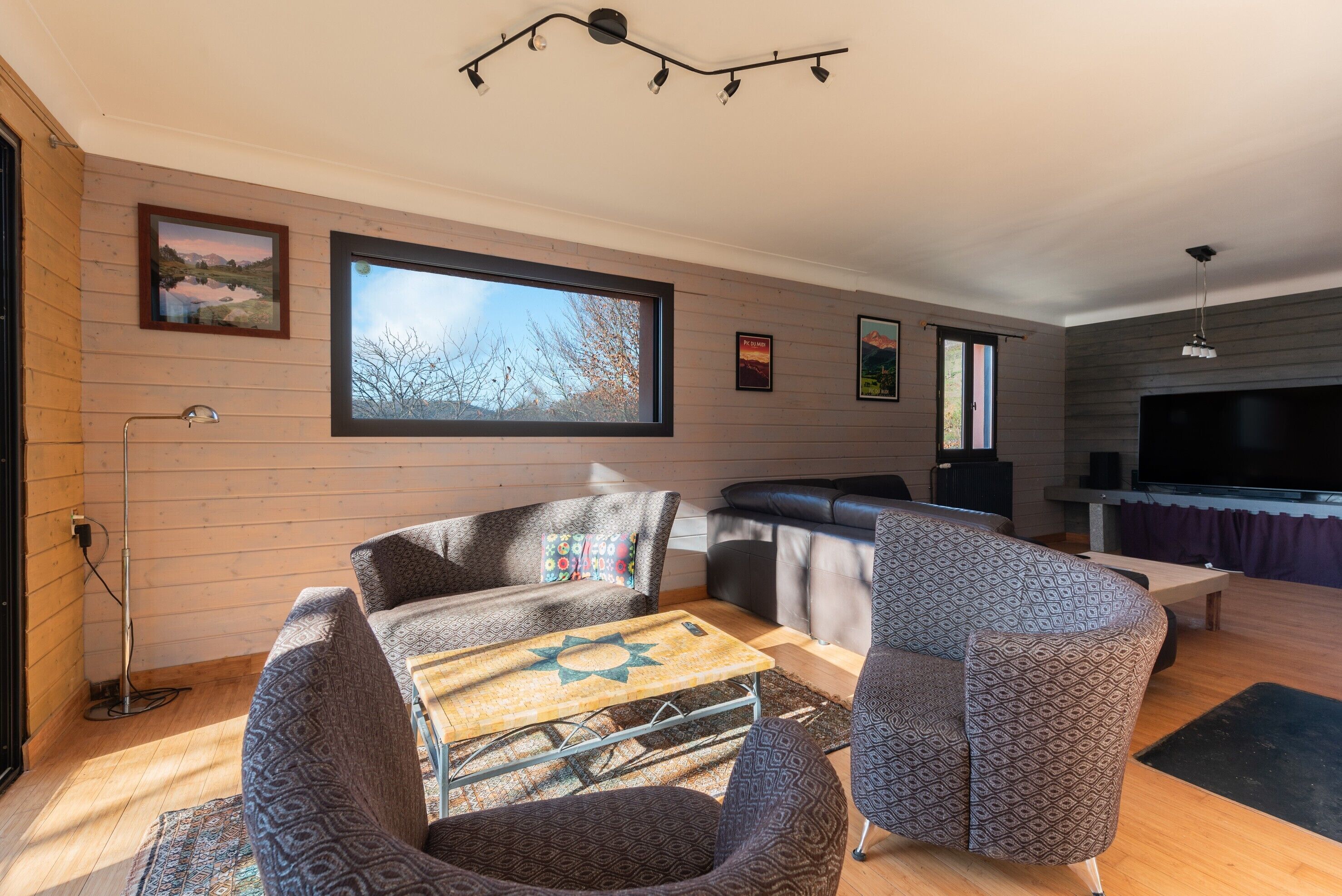 A modern living room with a large window, a sofa, a patterned armchair, a coffee table, and a flat-screen TV.