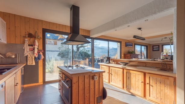 Une cuisine moderne avec des meubles en bois, un îlot central et de grandes fenêtres offrant une vue sur les montagnes.