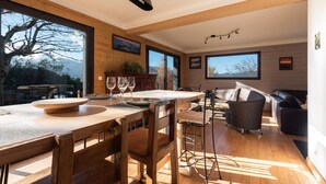 Un espace de vie moderne comprenant une table à manger en bois, des tabourets de bar, un canapé et de grandes fenêtres offrant une vue sur l’extérieur.