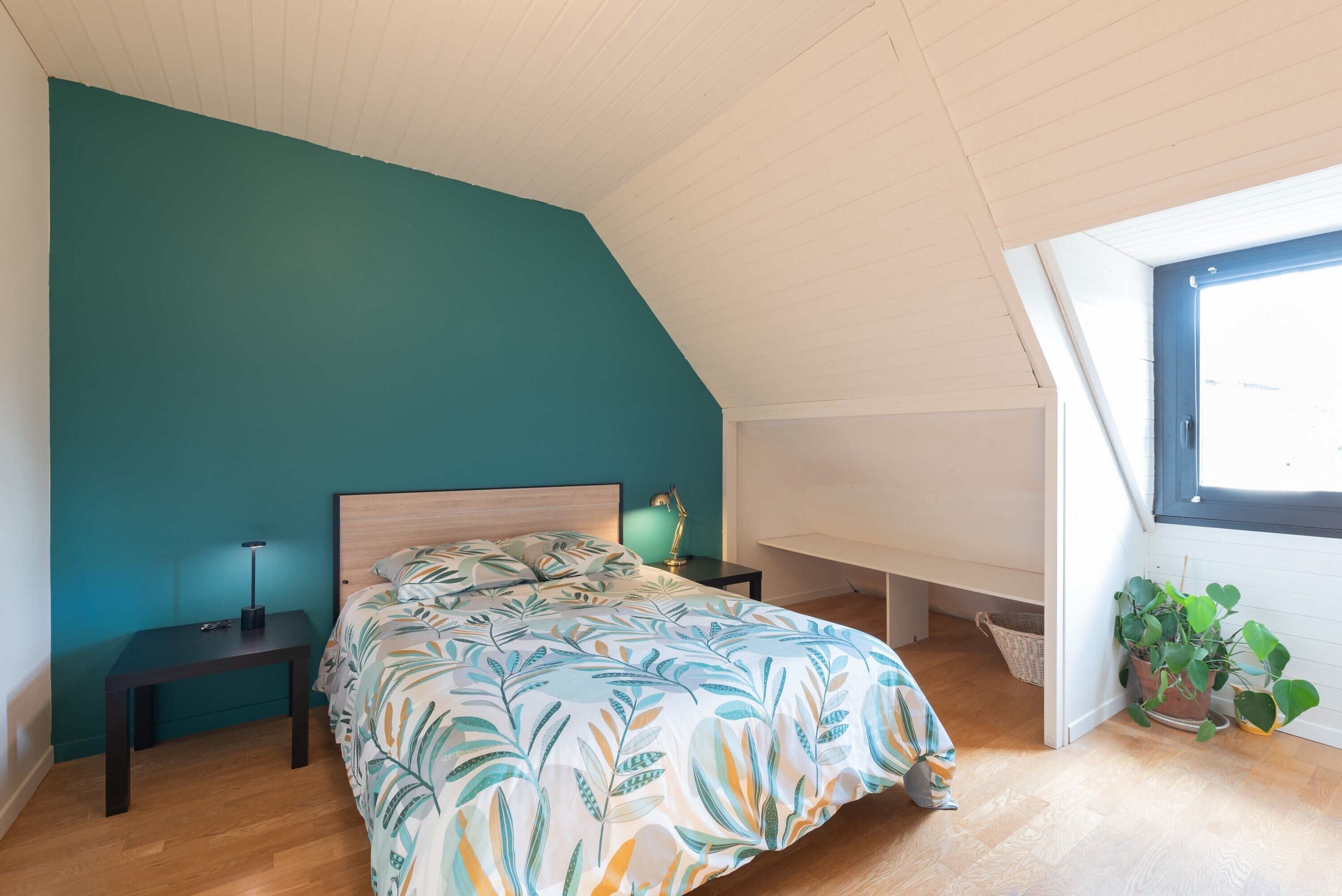 A bedroom with a bed, bedside tables, a nightstand, a window, and a potted plant.