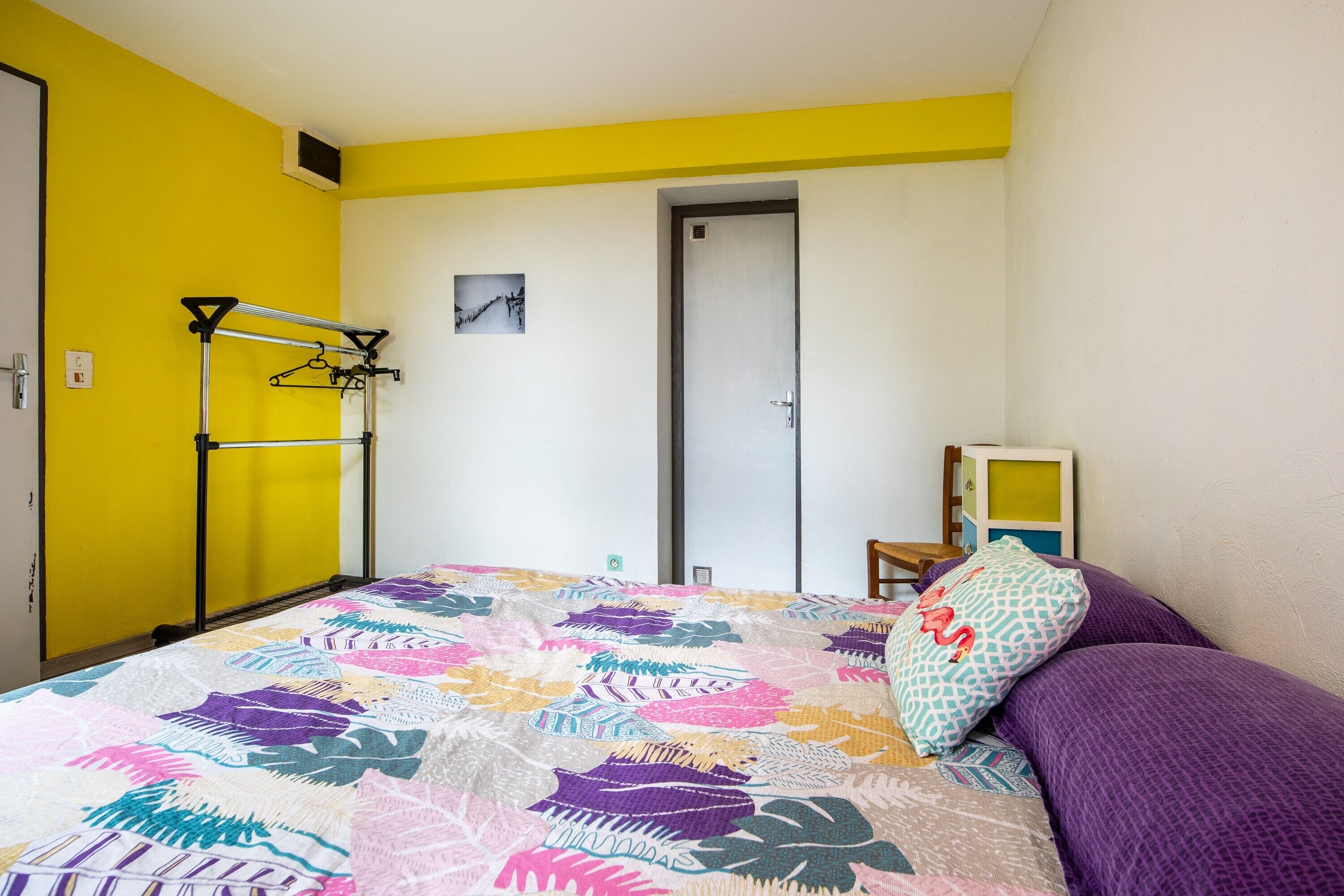 A bedroom with a bed, a door, a wall-mounted drying rack, and a small table with a chair.