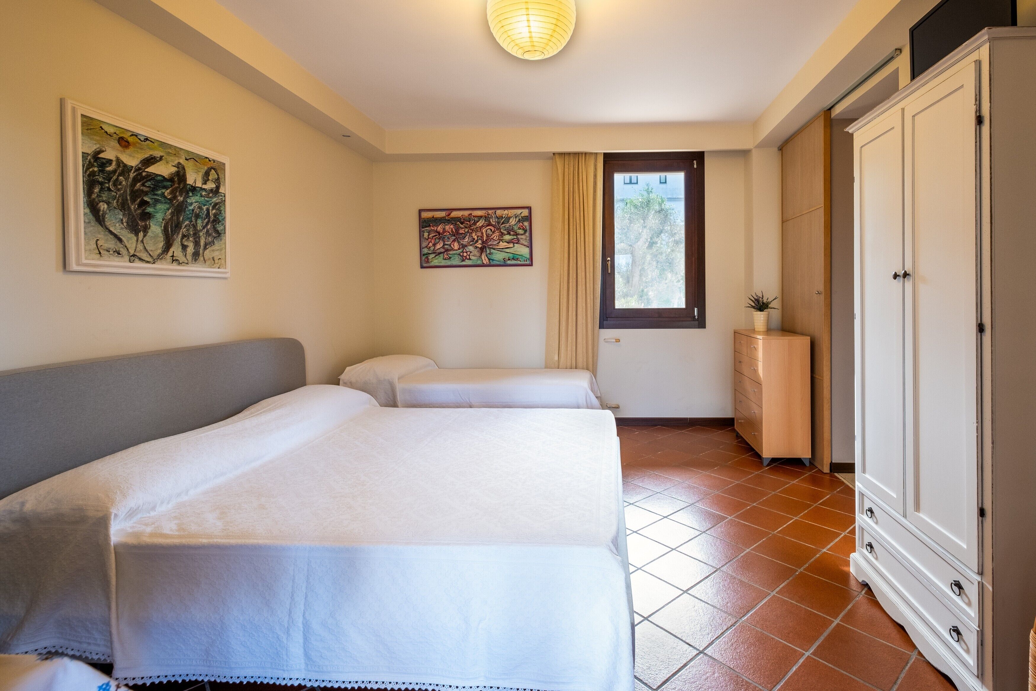 Ein Hotelzimmer mit zwei Betten, einem Kleiderschrank, einem Fenster mit Vorhängen und Bildern an den Wänden.