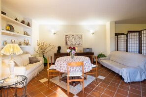 A living room with a sofa, a dining table with chairs, a lamp, and shelves with decorative items.