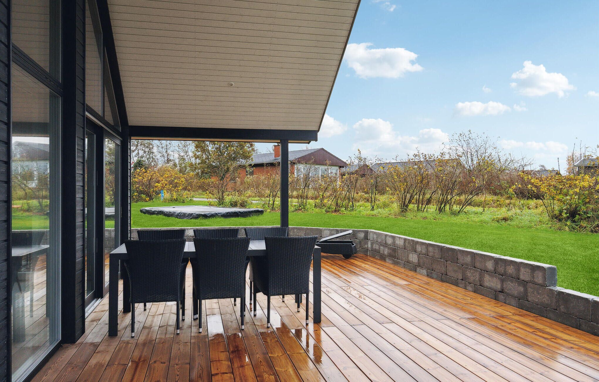 Um pátio exterior com deck de madeira, área de estar coberta com cadeiras e vista para um jardim e casas ao fundo.