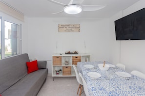 Een kamer met een grijze bank, een eettafel gedekt voor vier personen, een plafondventilator, een televisie aan de muur en een welkomstbord.