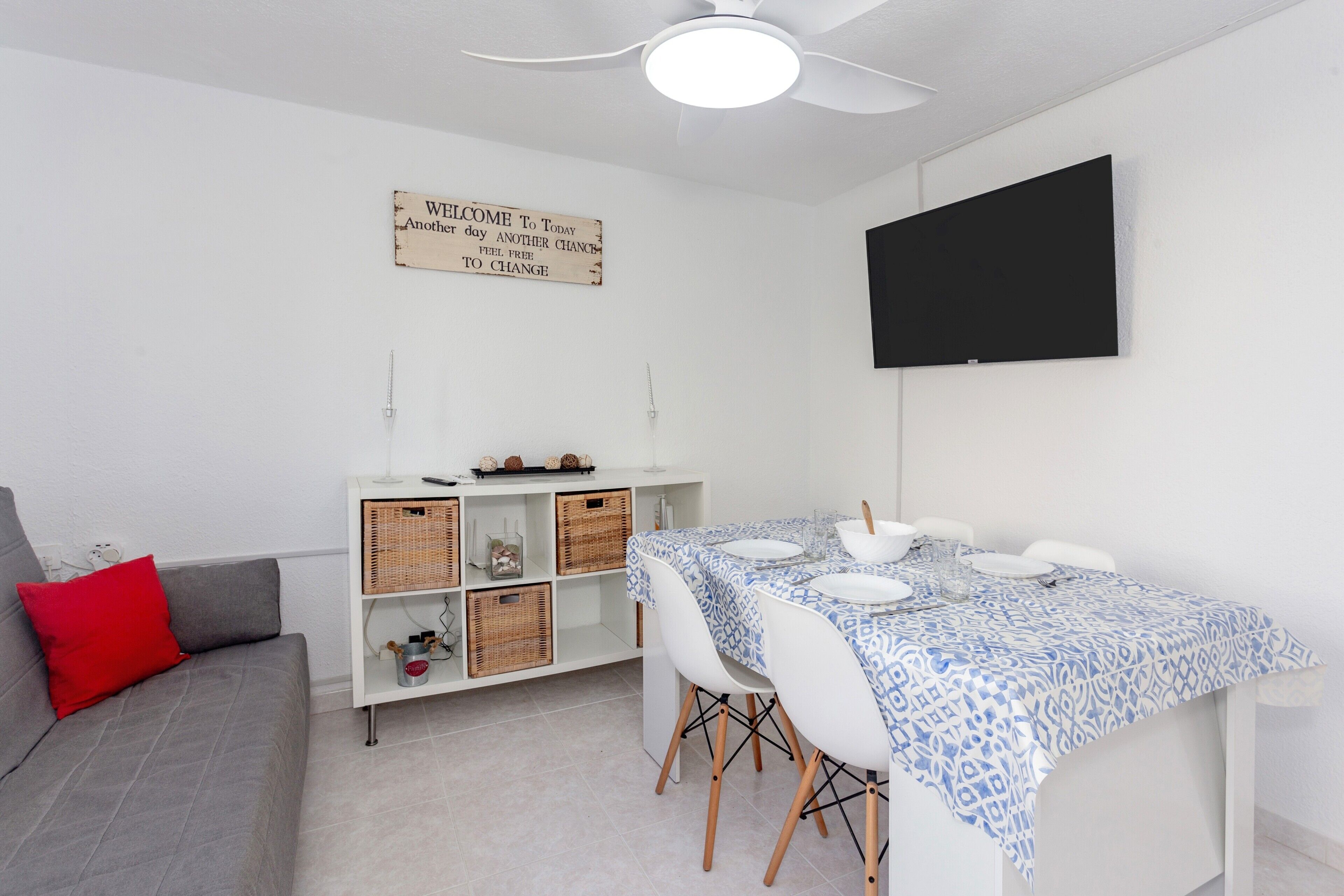 Un coin repas avec une table dressée pour quatre personnes, un canapé avec un coussin rouge, un meuble blanc avec des paniers et un ventilateur de plafond.