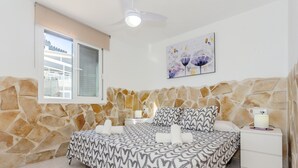 A bedroom with a stone wall, a bed with a patterned bedspread, a bedside table with a lamp, and a window with blinds. - Cala En Forcat Apartment with Shared Pool, Communal Terrace, and Wi-Fi (Menorca)