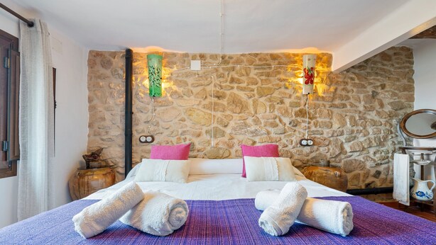 A bedroom with a stone wall, a bed with white linens, two pillows, a purple blanket, and two rolled towels.