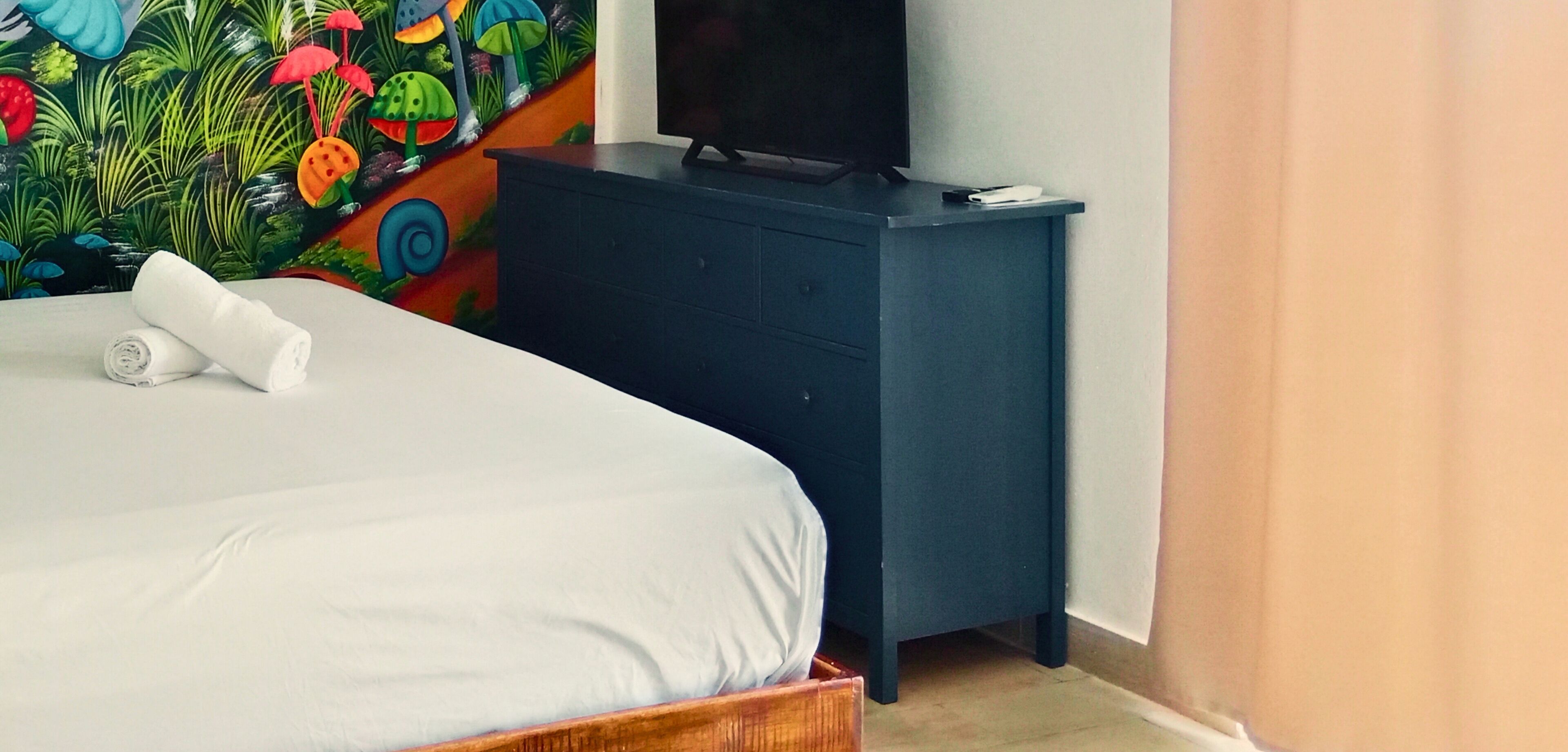 A neatly made bed with a white comforter and a rolled white towel, a dark blue dresser with a flat-screen TV on top, and a colorful mural on the wall behind the dresser.