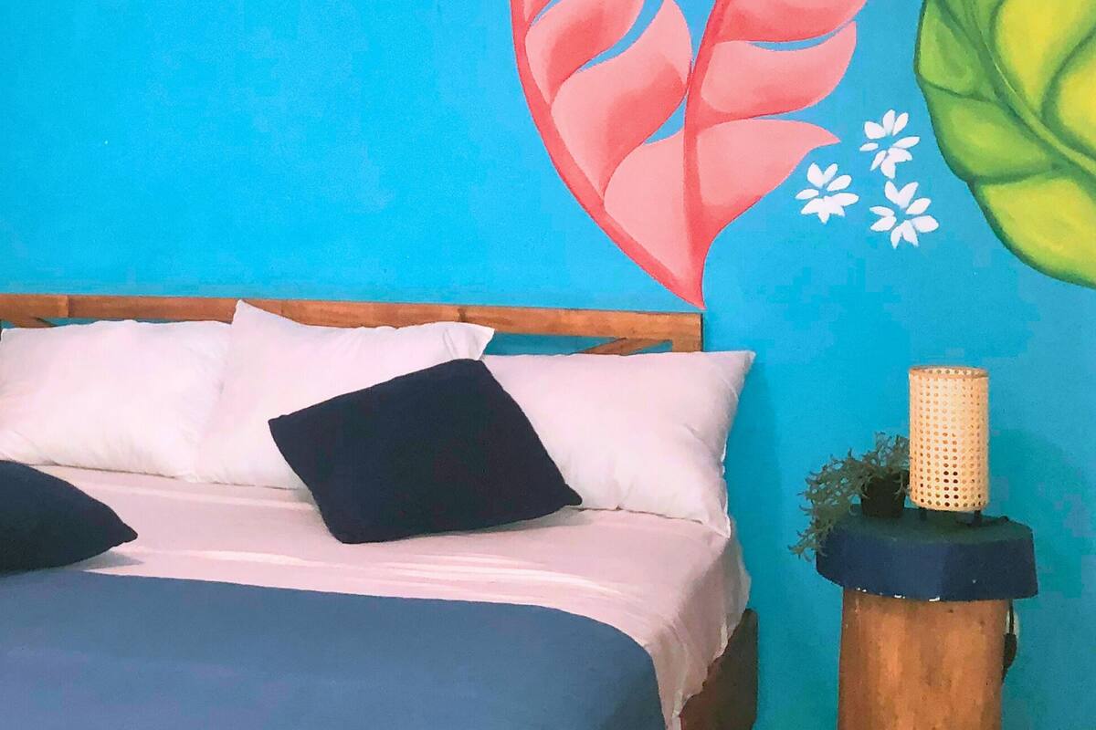Una cama con almohadas blancas y negras, una colcha azul y un cabecero de madera. Hay dos mesitas de noche con cestas tejidas, una con un certificado y la otra con una planta.