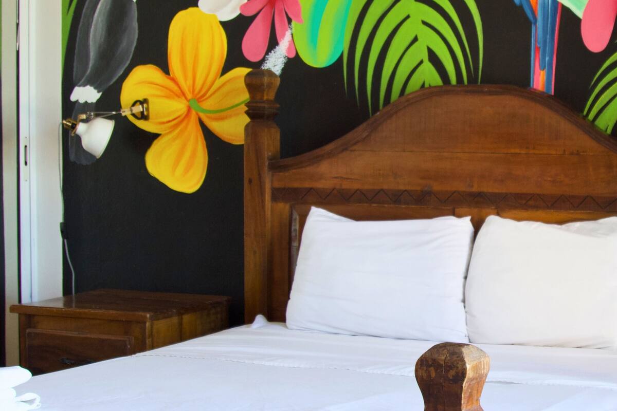 Una cama con cabecera de madera, ropa de cama blanca y una pared decorada con motivos florales tropicales.