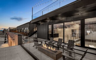 Un área de estar exterior moderna con una chimenea, barandillas de vidrio y una escalera que conduce a un nivel superior.
