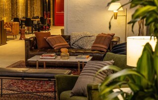 Una sala de estar acogedora con un sofá de cuero, un sillón verde y una mesa de centro con libros. Se ve una puerta roja al fondo y una lámpara en la pared.