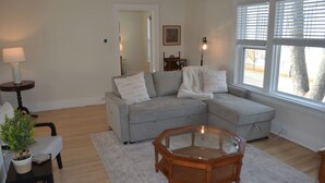 A living room with a grey sectional sofa, a glass coffee table, a small side table with a lamp, and a view through a window with blinds.