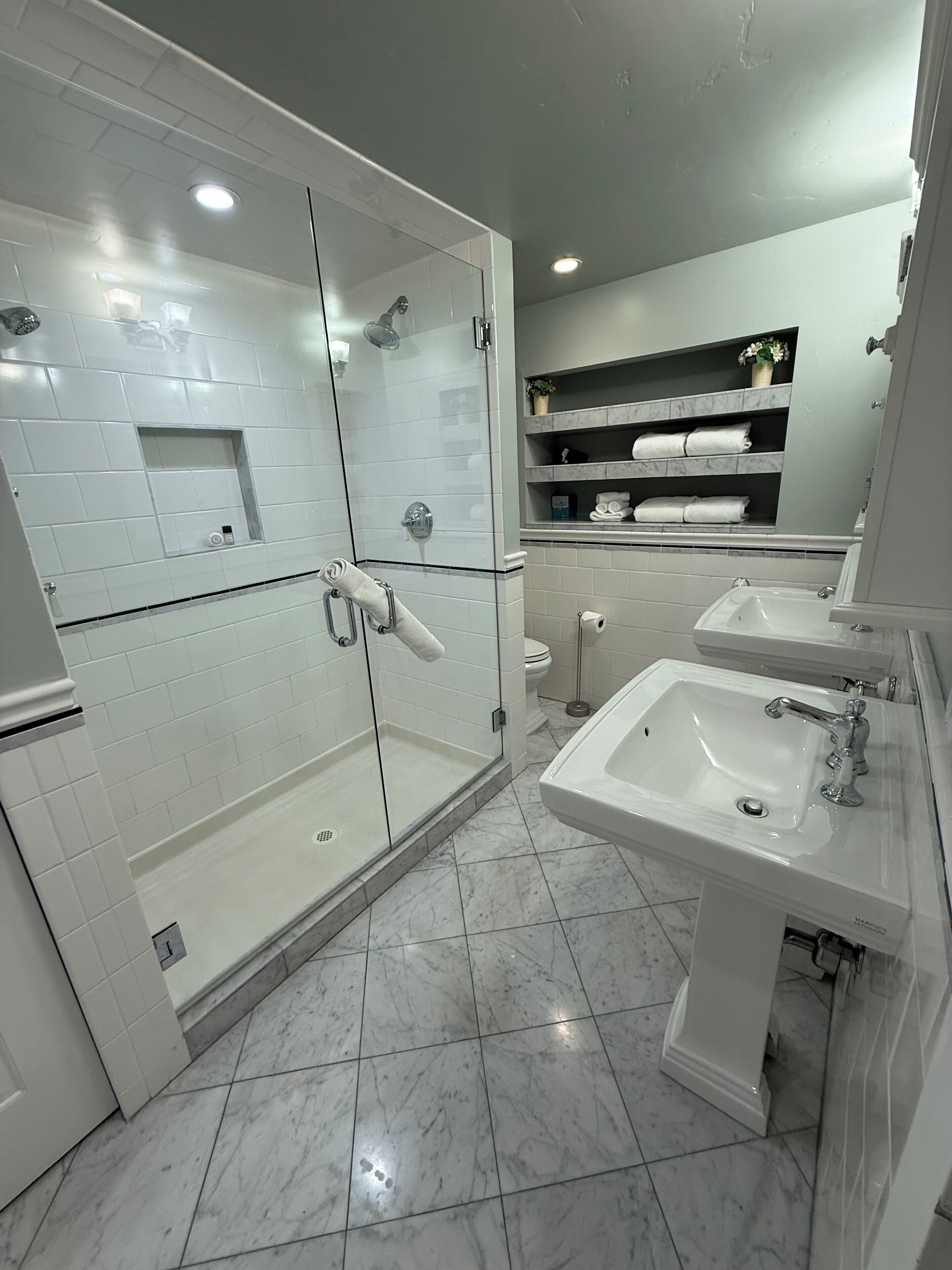 Une salle de bain moderne avec une douche à parois de verre, des murs recouverts de carreaux blancs, un sol en marbre et deux lavabos avec miroirs.