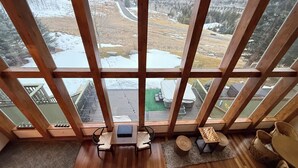 Ein Zimmer mit großen Fenstern, das einen Blick auf eine verschneite Landschaft und Berge bietet. Im Inneren befindet sich ein Holzboden mit Gartenmöbeln.