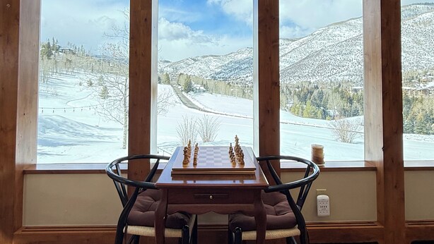 Ein Holztisch mit Schachfiguren vor großen Fenstern mit Blick auf eine verschneite Landschaft.