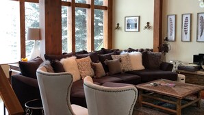 Ein gemütliches Wohnzimmer mit einem dunkelbraunen Sofa, zwei weißen Sesseln, einem Holztisch und einem großen Fenster mit Blick auf schneebedeckte Bäume.