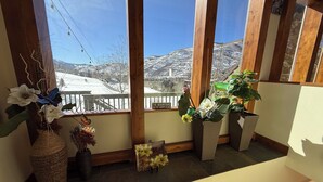 Ein Zimmer mit großen Fenstern mit Blick auf eine verschneite Berglandschaft. Mehrere Kübelpflanzen und ein dekoratives Schild stehen auf der Fensterbank.