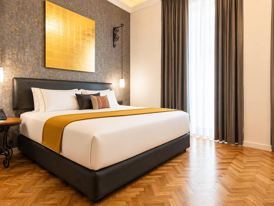 A hotel room with a large bed, a bedside table with a telephone, a wall-mounted artwork, and a window with curtains.